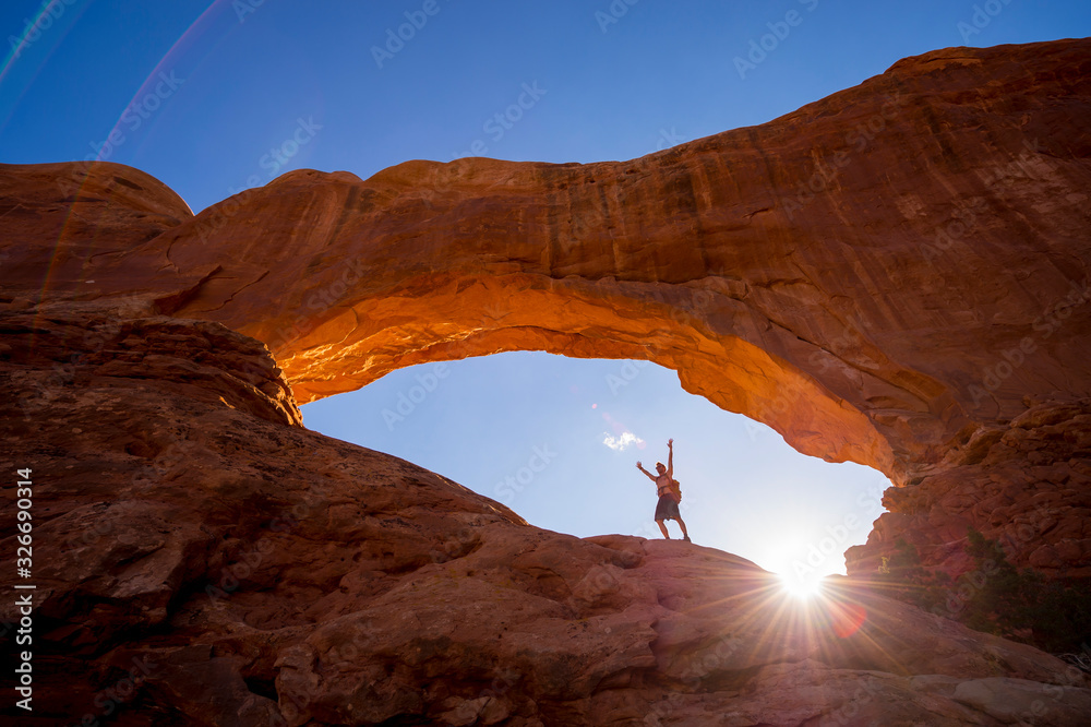 A hiker raising his arms to the sky standing under a dramatic natural rock arch in front of golden sun