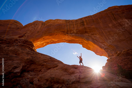 A hiker raising his arms to the sky standing under a dramatic natural rock arch in front of golden sun