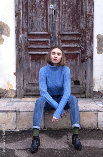 Beautiful young girl is sitting on the stair in the blue jeans and blue sweater near old brown door. Street photo session. Street fashion.
