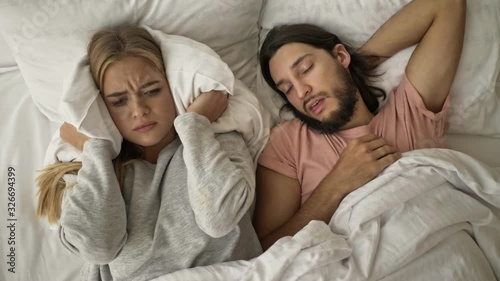 A displeased disappointed young woman is covering her ears with a pillow while her boyfriend is snoring in the bed at the bedroom photo