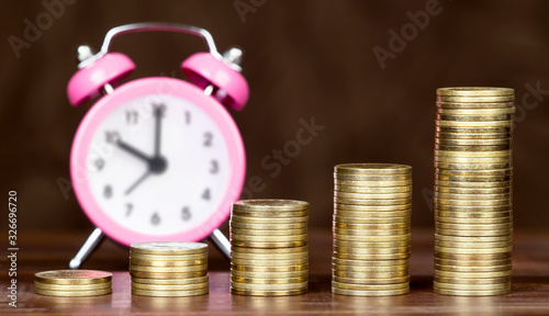 Financial freedom concept. Gold money coins stack with pink alarm clock on brown background.