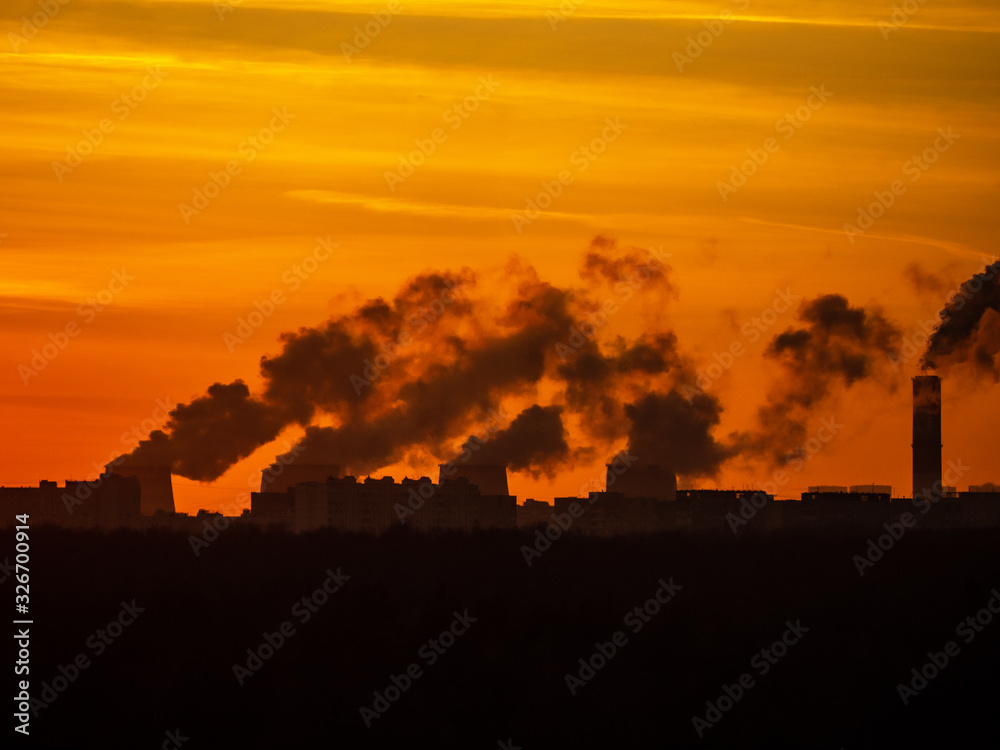 Cityscape silhouette with clouds of smoke from smoking chimneys of thermal power station and pipes of the industrial plant and cloudy orange sunset sky