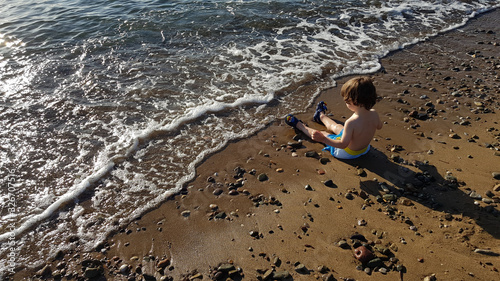 Four year old boy sitting by the sea at sunset, playing with water