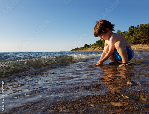 Four year old boy sitting by the sea at sunset, playing with water