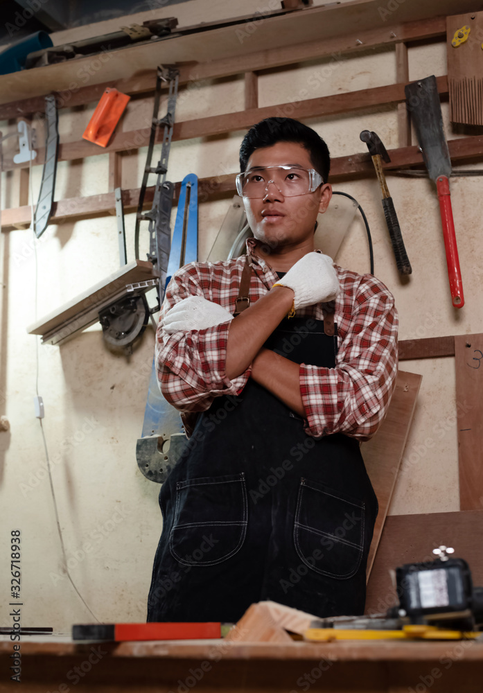 Handsome man wearing blue apron and protective glasses,standing beside tools and equipment for wood work on wall,at home studio handmade,,blurry light around