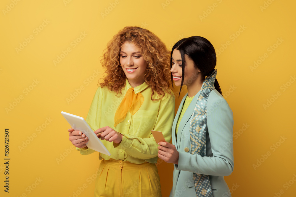 Fototapeta premium happy redhead girl using digital tablet while shopping online with african american woman holding credit card on yellow
