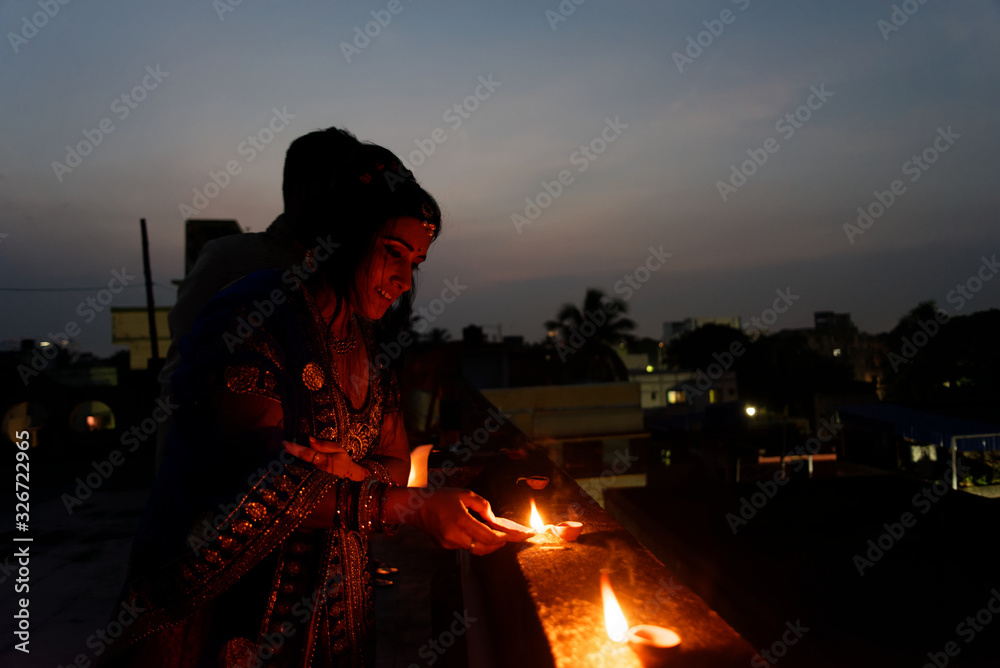 Obraz premium Young and beautiful Indian Gujarati couple in Indian traditional dress lightening Diwali diya/lamps sitting on the terrace in blue hour on Diwali evening. Indian lifestyle and Diwali celebration