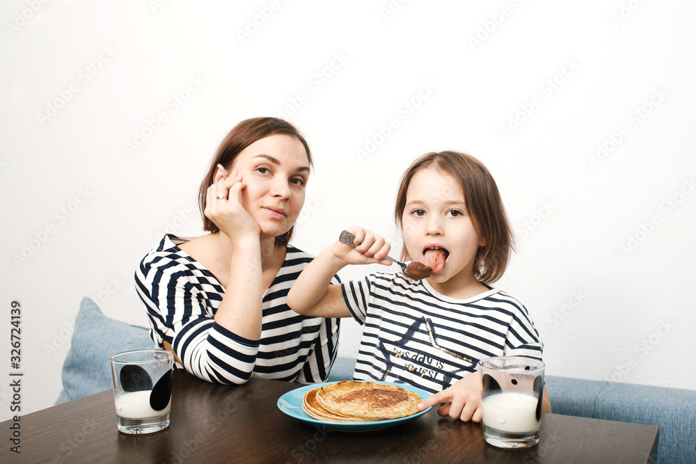 Mother and little daughter eating pancakes with milk in the morning in light kitchen with white background Mother and little daughter eating pancakes with milk in the morning in light kitchen with white background