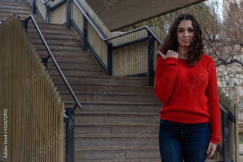Portrait of attractive young woman model advertising product with pointing finger aside on copyspace isolated, in the park, orange sweater and jeans, long curly hair. Place for your text in copy space