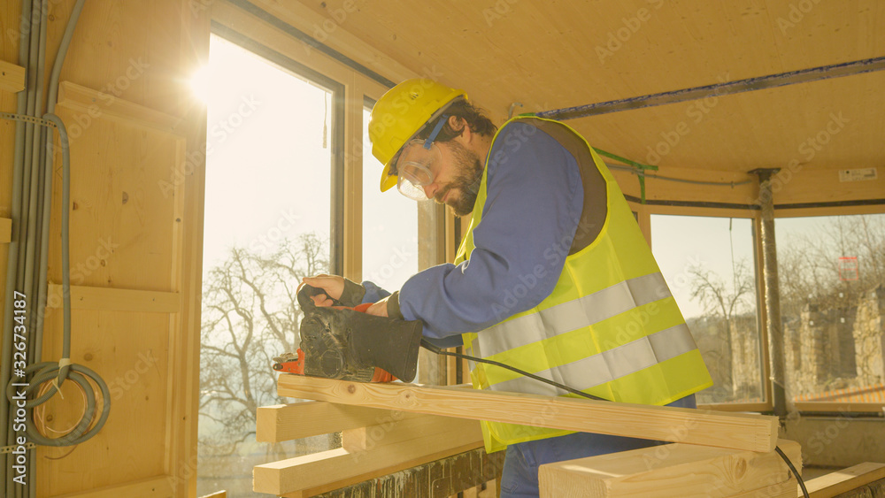 CLOSE UP: Young man working on a prefabricated house buffs a plank with a sander