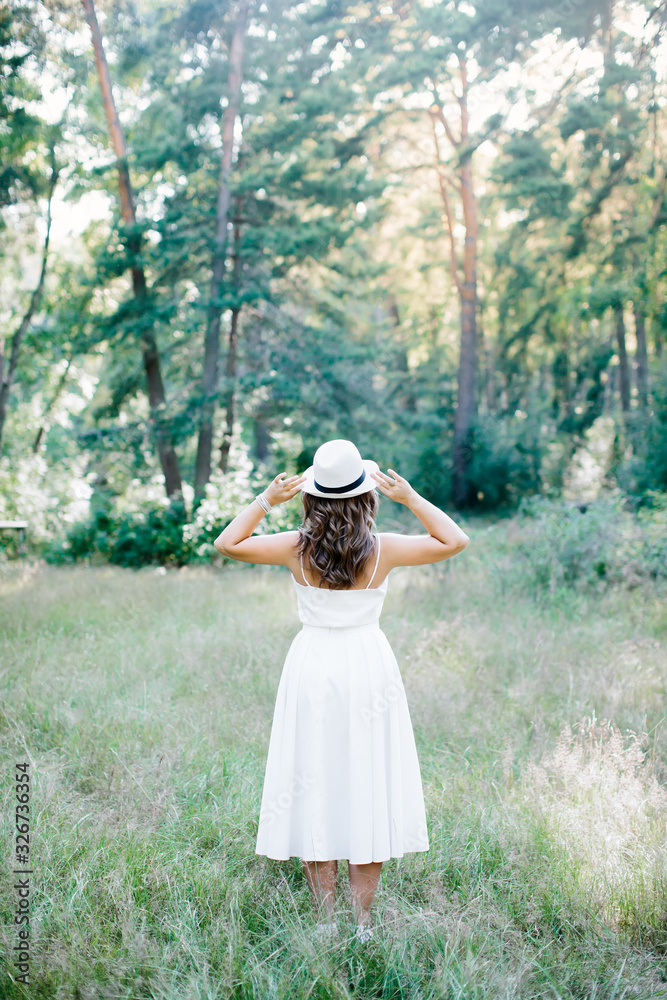 Girl in white clothes and a hat stands with his back on nature.