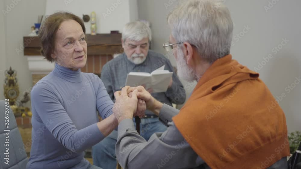 Portrait of positive senior Caucasian woman talking with mature man and laughing. Elderly couple flirting as lonely man reading at the background in nursing home. Portrait of positive senior Caucasian woman talking with mature man and laughing. Elderly couple flirting as lonely man reading at the background in nursing home.