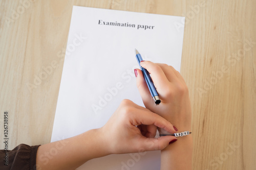 Student girl hand with bracelet and love word. Empty examination paper sheet on the table. First love during university exam. Concept