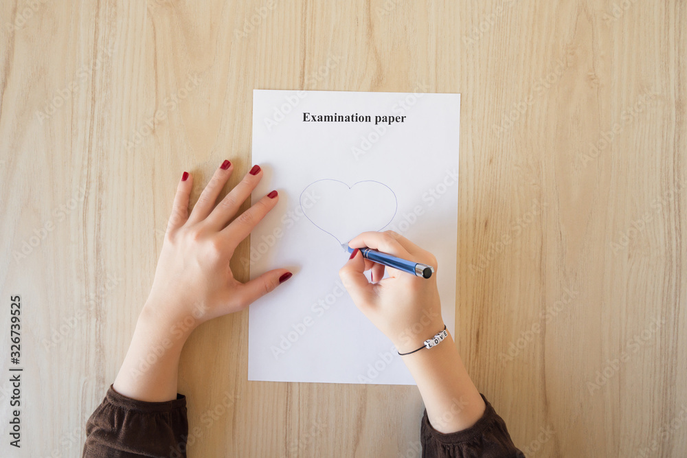 Student girl hand with bracelet and love word drawing a heart on empty examination paper sheet on the school desk. First love during university exam. Concept