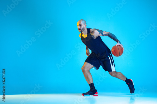 Young basketball player of team wearing sportwear training, practicing in action, motion isolated on blue background in neon light. Concept of sport, movement, energy and dynamic, healthy lifestyle.