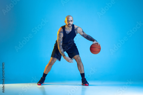 Young basketball player of team wearing sportwear training, practicing in action, motion isolated on blue background in neon light. Concept of sport, movement, energy and dynamic, healthy lifestyle.