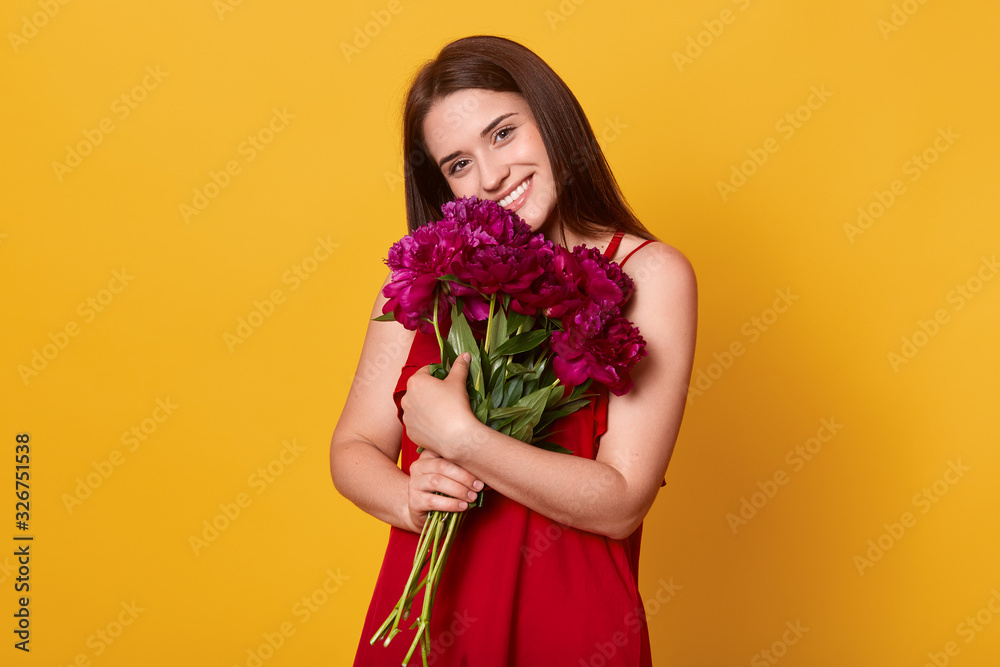 Horizontal shot of happy woman hugs big bunch with flowers, posing isoalted over yellow background, lady wearing red sundress looking directly at camera with charming smile, brunette girl with bouquet Horizontal shot of happy woman hugs big bunch with flowers, posing isoalted over yellow background, lady wearing red sundress looking directly at camera with charming smile, brunette girl with bouquet