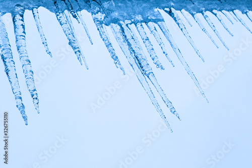 Big blue icicles under the roof against the background of a bright sky. Thaw and warming winter weather. Roof icing. Ice frost under the roof of the house. Frosty weather.