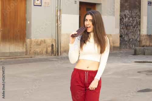 Portrait of happy attractive caucasian young woman model eating chocolate, lovely smiling, in the city, white top and red pants, dark air. Place for your text in copy space.