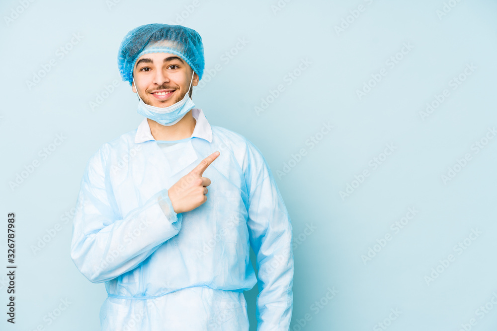 Young arabian surgeon man isolated against on a blue background smiling and pointing aside, showing something at blank space. Young arabian surgeon man isolated against on a blue background smiling and pointing aside, showing something at blank space.