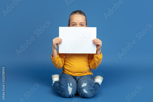 Portrait of sad little girl sitting isolated on blue studio background. How it feels to be autist. Modern problems, new vision of social issues. Concept of autism, childhood, healthcare, medicine. photo