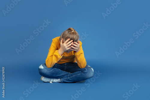 Portrait of sad little girl sitting isolated on blue studio background. How it feels to be autist. Modern problems, new vision of social issues. Concept of autism, childhood, healthcare, medicine. photo