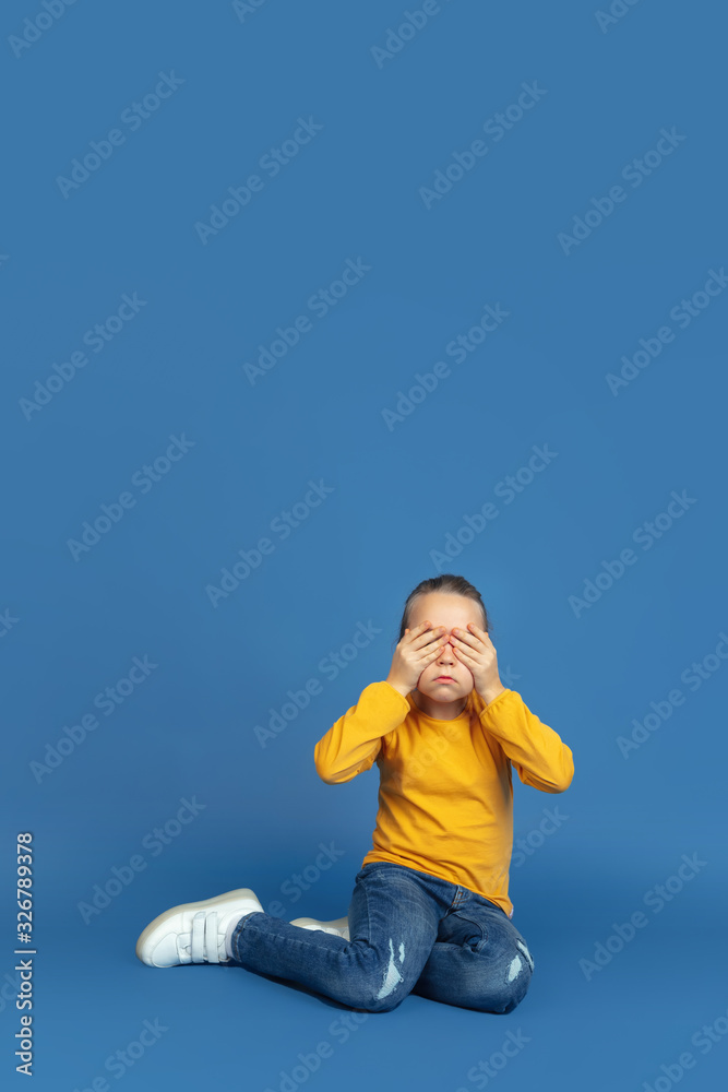 Portrait of sad little girl sitting isolated on blue studio background. How it feels to be autist. Modern problems, new vision of social issues. Concept of autism, childhood, healthcare, medicine. Portrait of sad little girl sitting isolated on blue studio background. How it feels to be autist. Modern problems, new vision of social issues. Concept of autism, childhood, healthcare, medicine.