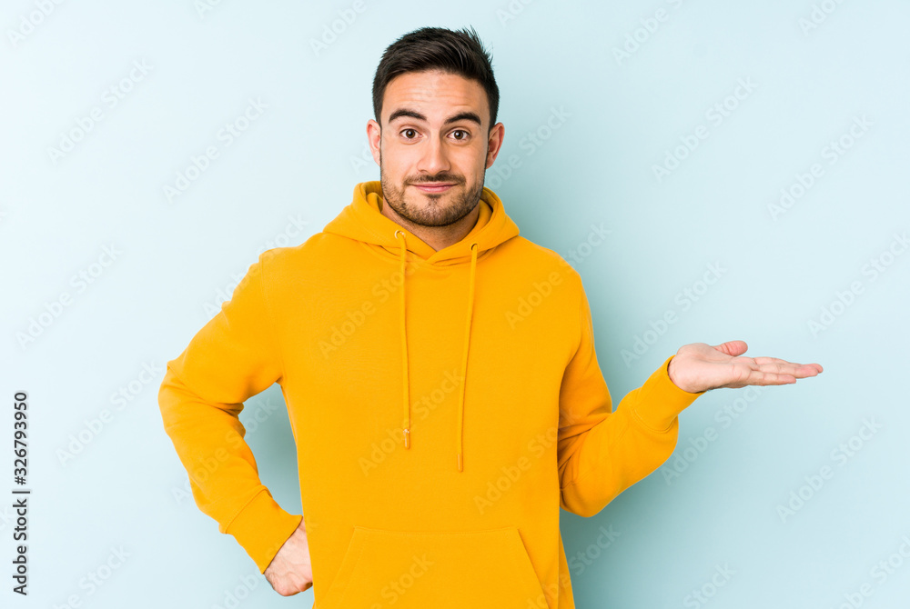 Young caucasian man isolated on blue background showing a copy space on a palm and holding another hand on waist. Young caucasian man isolated on blue background showing a copy space on a palm and holding another hand on waist.