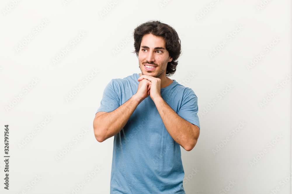Young handsome man against a white background keeps hands under chin, is looking happily aside.