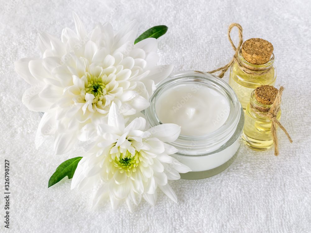 Face cream in an open glass jar, two bottles of chrysanthemum essential oil and two white chrysanthemums on a white terry towel. Beauty, skincare and cosmetology. Face cream in an open glass jar, two bottles of chrysanthemum essential oil and two white chrysanthemums on a white terry towel. Beauty, skincare and cosmetology.
