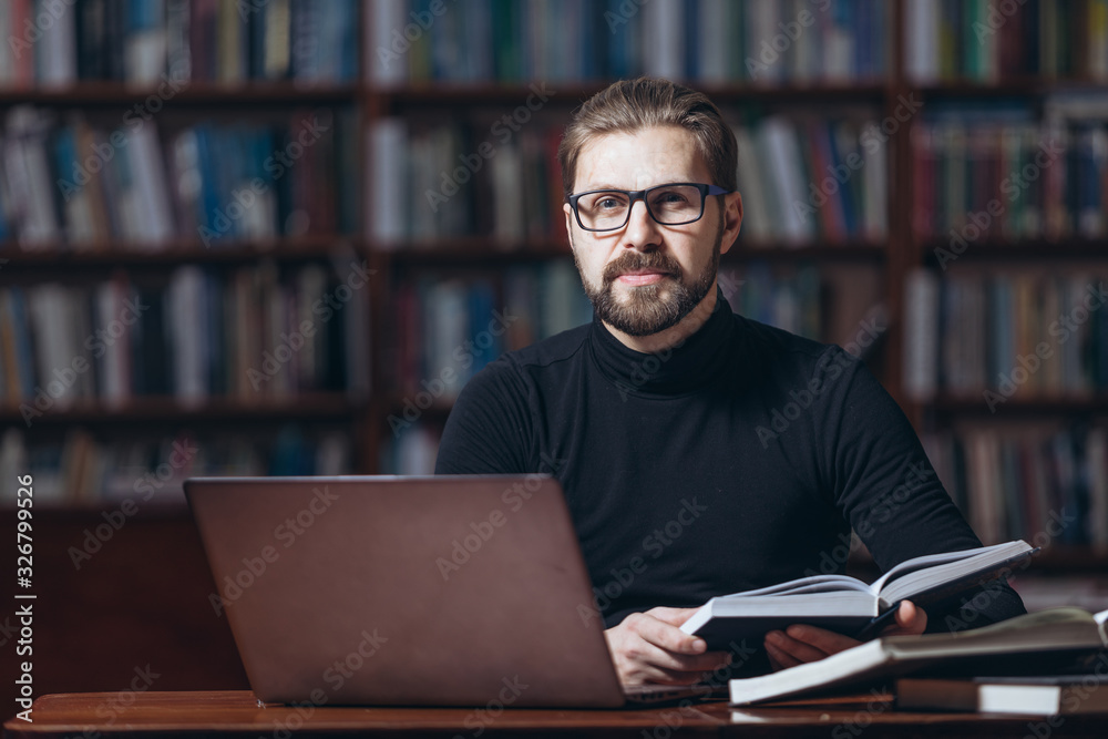 Confident bearded man in black polo neck sitting at table with open book and laptop and looking at camera. Mature male in eyeglasses working at public library