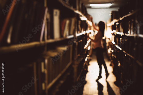 Blur background of young lady with long hair in casual clothing trying to get book from the highest shelf at dark bookstore. Beautiful woman choosing literature for gaining new knowledge.