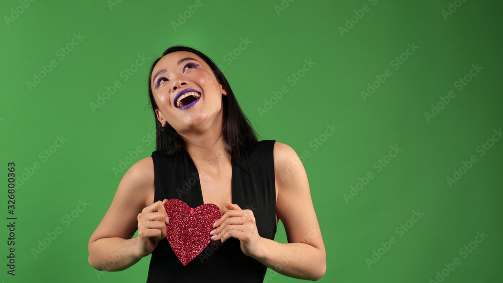 Asian smiling woman dancing with a paper red heart in her hands and covering her face with it on an isolated green background, concept of the holiday of lovers