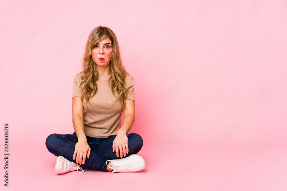 Young blonde caucasian woman sitting on a pink studio shrugs shoulders and open eyes confused. Young blonde caucasian woman sitting on a pink studio shrugs shoulders and open eyes confused.