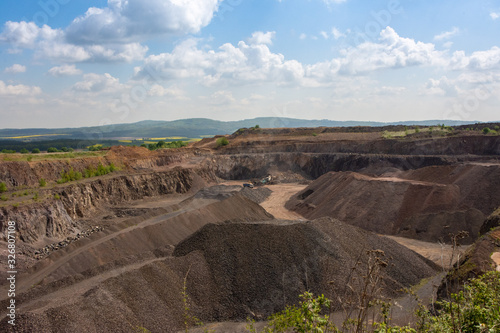 View of basalt (brown rock) quarry with rock exploitation during summer - blue sky with clouds, open-pit plant and village and background