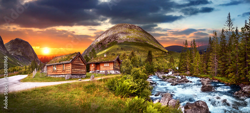 Beautiful magic forest with two wooden houses near blue wild river in the sunset view. Amazing evening norway country panomama.