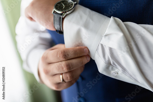 A man in a white shirt and blue vest is buttoning his cuffs. A stylishly dressed businessman adjusts his cuffs