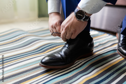 Man ties his shoelaces on black expensive classic shoes in the room. Businessman in a blue suit and patent leather shoes.
