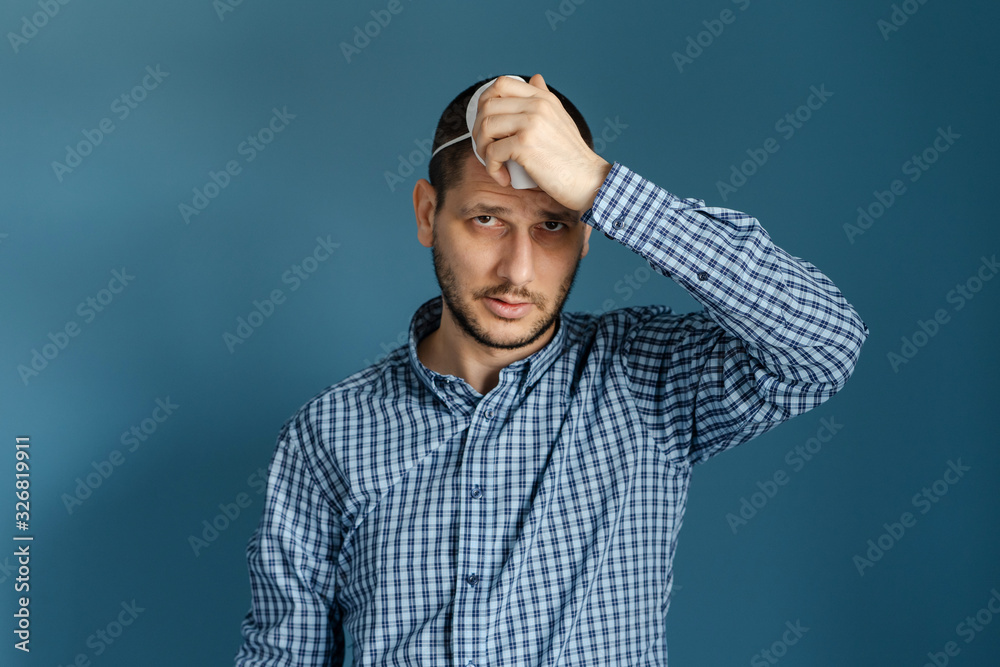 Portrait of adult caucasian man standing in front of the blue wall wearing shirt holding protective mask on his face against virus flue bacteria protection health issues front view putting on Portrait of adult caucasian man standing in front of the blue wall wearing shirt holding protective mask on his face against virus flue bacteria protection health issues front view putting on