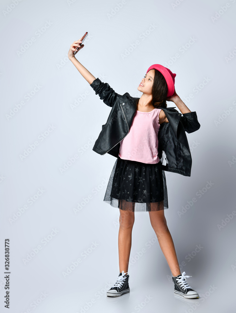 Teenage kid in black leather jacket, skirt and pink hat. She taking selfie using smartphone, posing isolated on white. Close up Teenage kid in black leather jacket, skirt and pink hat. She taking selfie using smartphone, posing isolated on white. Close up
