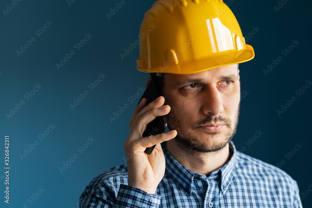 Close up portrait of young man engineer wearing yellow protecting safety helmet and shirt in front of blue wall isolated holding smart phone making a call talking Close up portrait of young man engineer wearing yellow protecting safety helmet and shirt in front of blue wall isolated holding smart phone making a call talking
