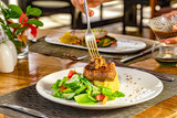 Grilled steak, coffee sauce, mashed potatoes and vegetable salad on a white plate, hand holding a fork and serving caramelized onions, blurred restaurant background