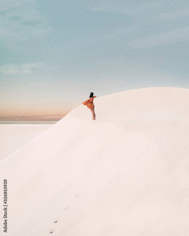A pretty young female traveler enjoying life in the desert. This artistic picture is taken in the dunes with a beautiful sunset as a background. The girl is wearing a dress that is moving by the wind.