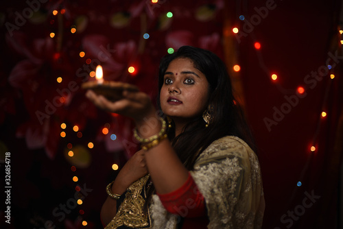 An young and beautiful Indian Bengali woman in Indian traditional dress is holding a Diwali diya/lamp in her hand standing in front of colorful bokeh lights. Indian lifestyle and Diwali celebration