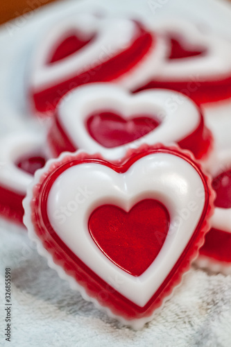 A red piece of handmade soap in the shape of a heart with a white insert  lying on a white towel against the background of five pieces of soap in the shape of a heart.