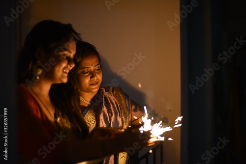 Two young and beautiful Indian Bengali women in Indian traditional dress are celebrating Diwali with diya/lamp and fire crackers on a balcony in darkness. Indian lifestyle and Diwali celebration