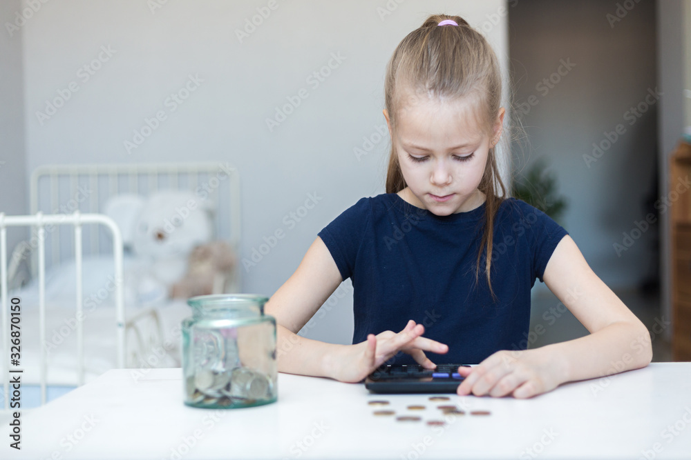 Cute little child girl counts on a calculator and puts coins in a glass bottle in her room. The child saves money on the concept of the future Cute little child girl counts on a calculator and puts coins in a glass bottle in her room. The child saves money on the concept of the future