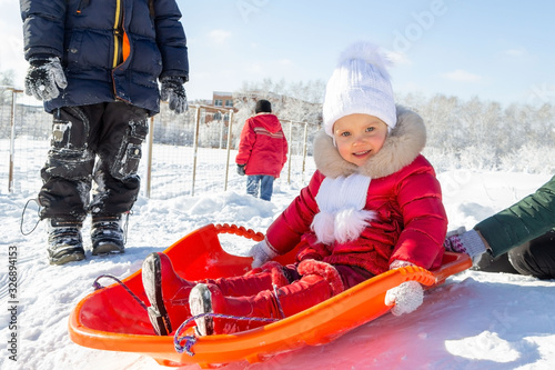 Winter activities for children. A little girl in a red coat sits on an orange sled before descending from a snow slide. Behind her are other children and a school in the distance. Snowy weather