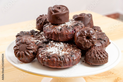 Delicious chocolate dietary candies and cookies for vegans made from natural ingredients. Tasty treats, sprinkled with coconut, on a white tray on wooden table. Healthy meal. Sweet and harmless.