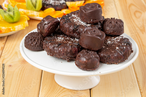 Tasty treats, sprinkled with coconut, on a white tray on wooden table. Delicious chocolate dietary candies and cookies for vegans made from natural ingredients. Healthy meal. Sliced fruits in back.
